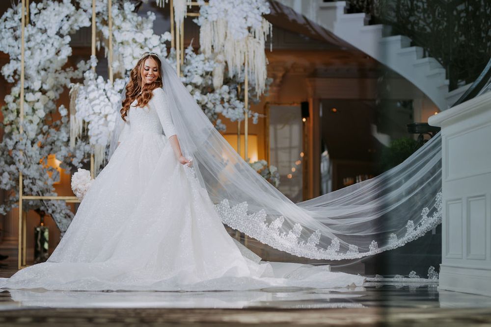 A bride in a flowing white ballgown poses gracefully in front of an elegant staircase with crystal chandeliers.