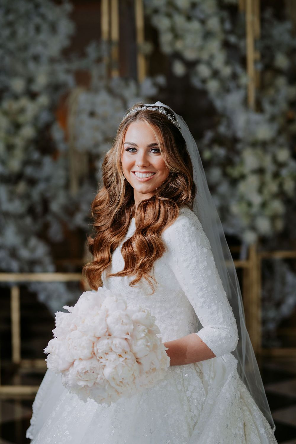 Bride in a stunning white wedding dress with long wavy hair and veil poses against a floral backdrop.