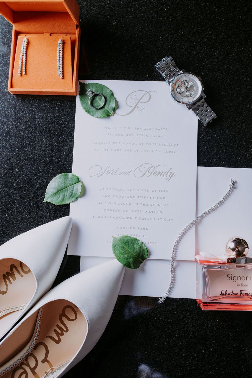 Wedding invitation suite displayed with decorative leaves and jewelry on a dark background.