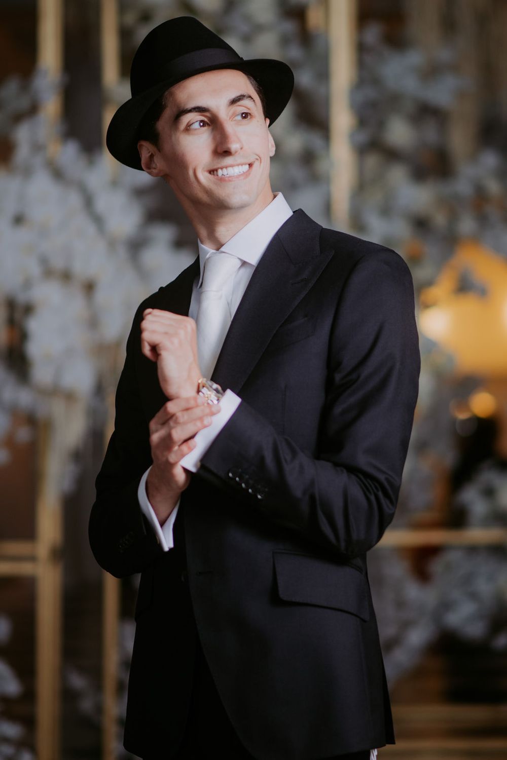 Groom in classic black tuxedo and top hat poses playfully against elegant backdrop.