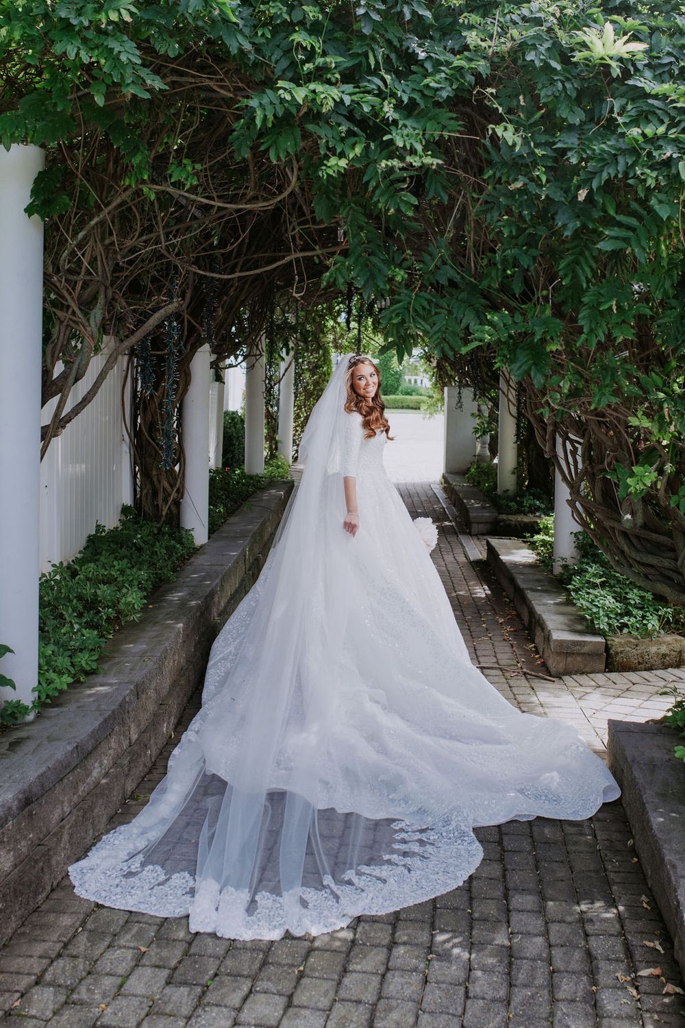 A bride in a flowing white wedding gown with a long train poses in a garden pathway lined with white columns and greenery.