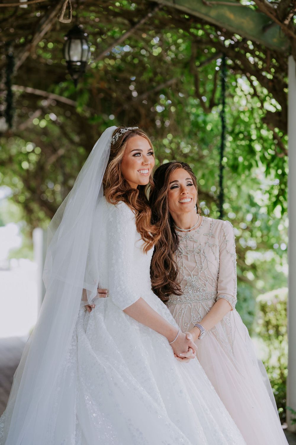 Two women in elegant white and cream dresses share a joyful moment under a garden archway with hanging lanterns.