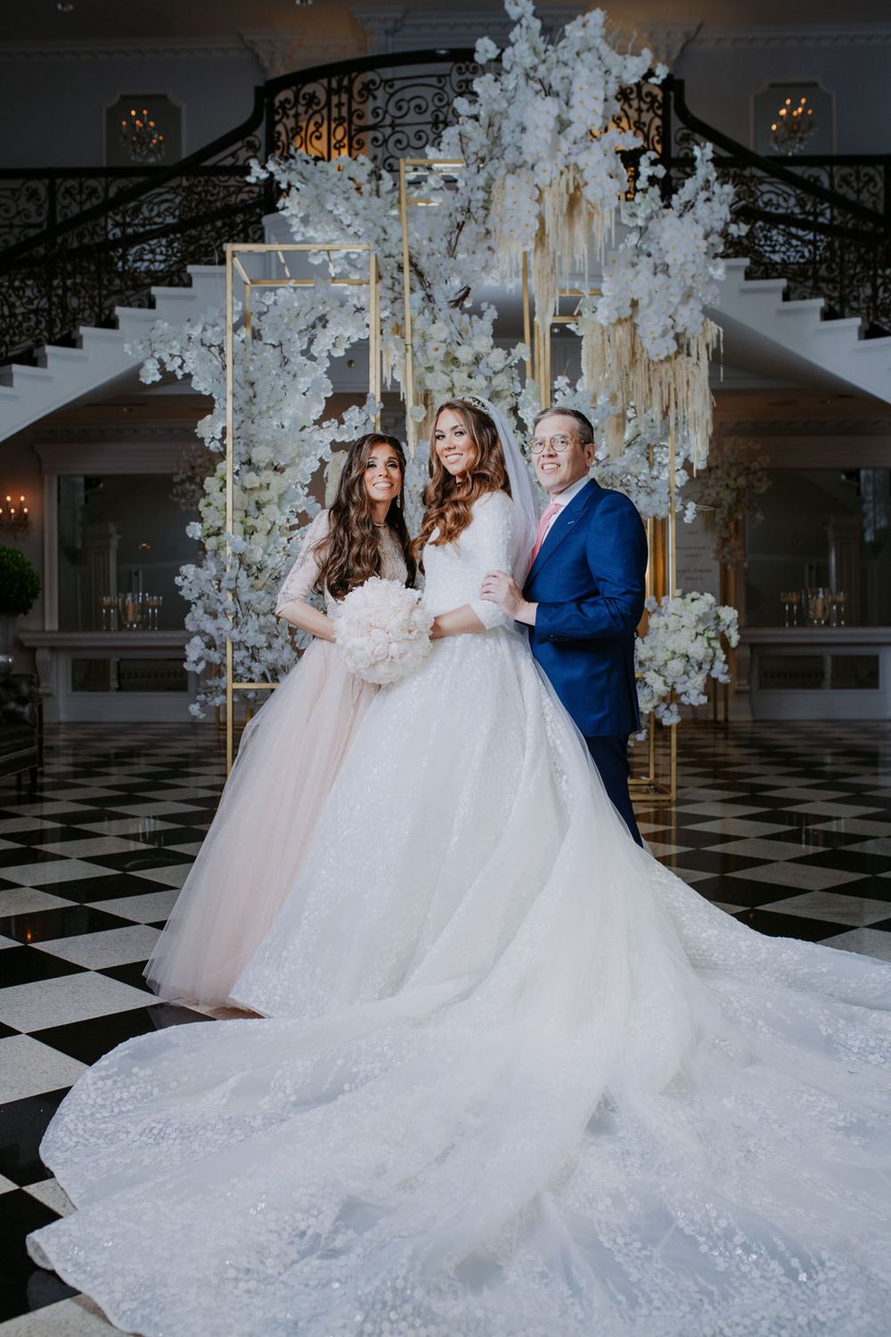 Wedding party in flowing white and blush gowns pose in grand ballroom with checkered floor.