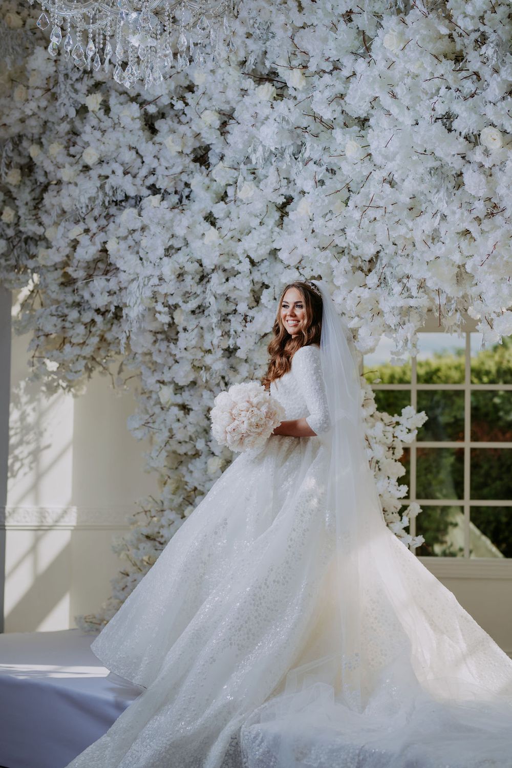 A stunning white wedding dress with a cathedral train against a backdrop of white floral decorations.