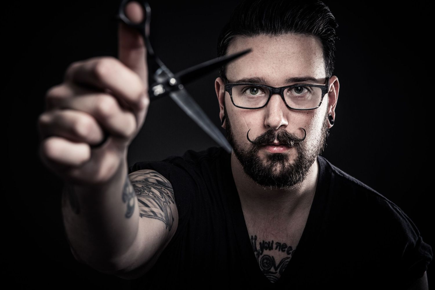 Professional portrait of someone wearing glasses holding scissors against dark background with dramatic lighting.