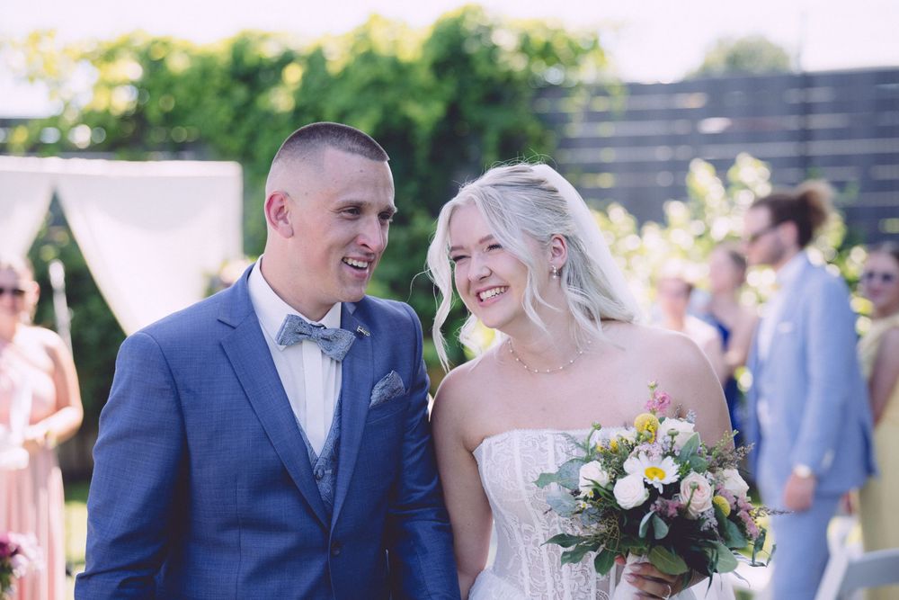 Happy newlyweds walk down the aisle, groom in blue suit, bride in strapless gown holding a floral bouquet.