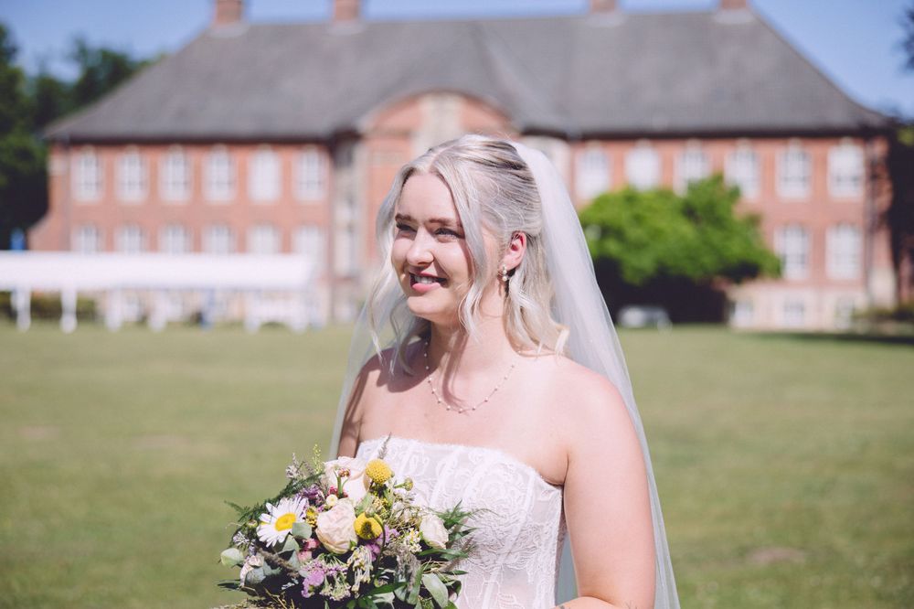 Bride in strapless lace gown holding wildflower bouquet smiling outdoors with red brick manor house in background.