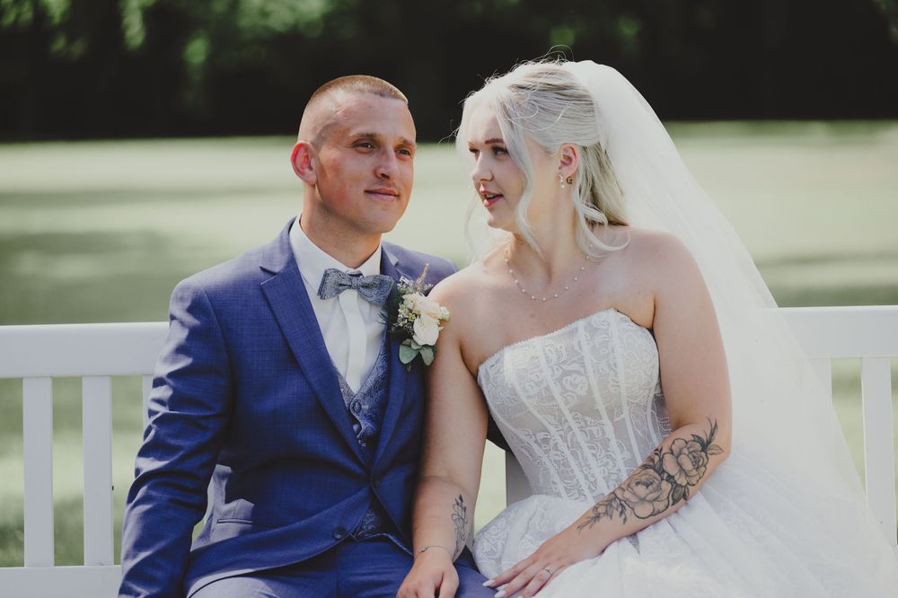 A bride in a white strapless gown and veil sits with a groom in a blue suit on a bench outdoors.