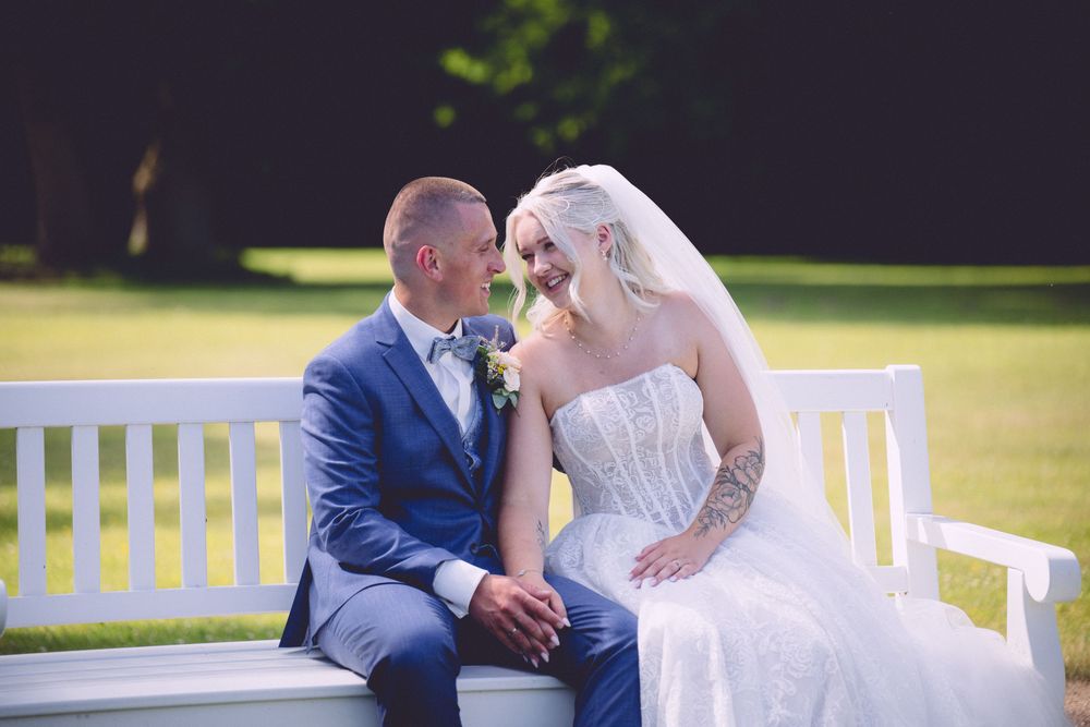 Bride and groom sitting together on a white bench in a sunny garden, sharing a romantic gaze on their wedding day.
