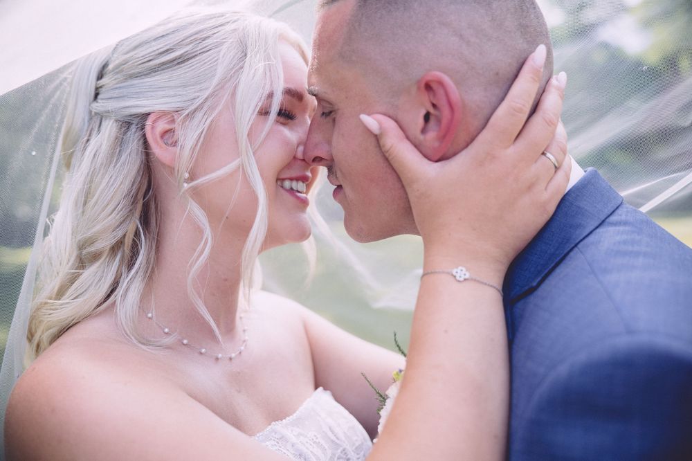 A bride and groom share a romantic moment under a veil, smiling and touching foreheads on their wedding day.