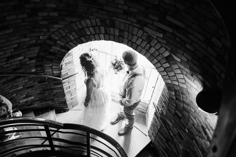 A dramatic black and white wedding photo taken through a brick archway tunnel with light at the end.
