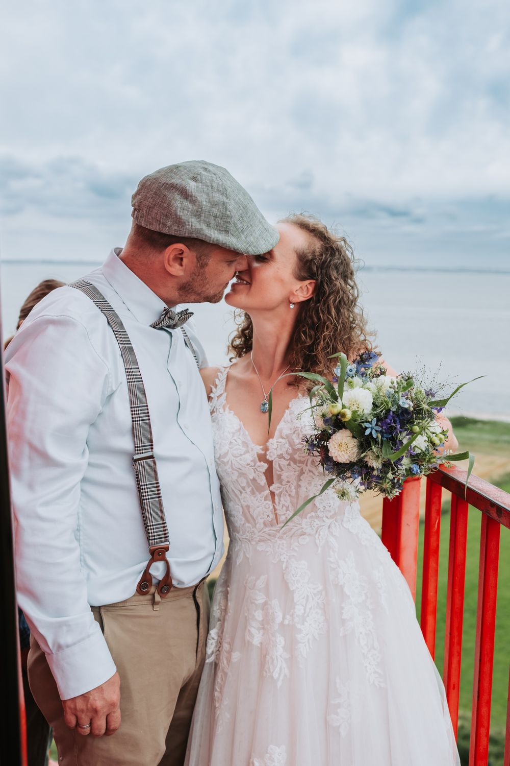 Two people share a romantic kiss at their waterfront wedding, with the person in a flowing lace dress holding a blue bouquet.