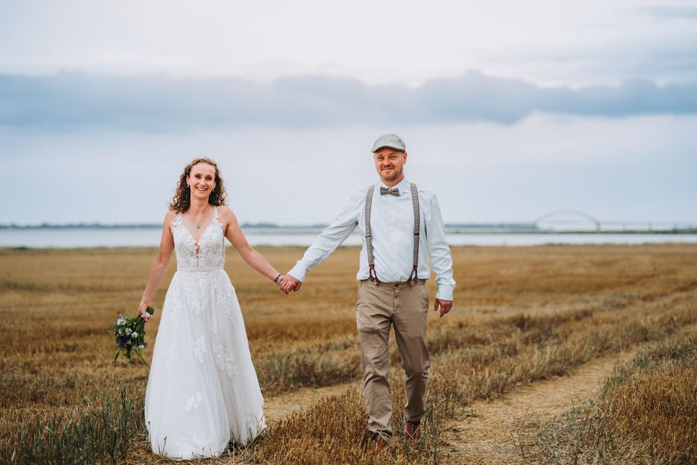 A couple in wedding attire holds hands while walking through a golden field at sunset.