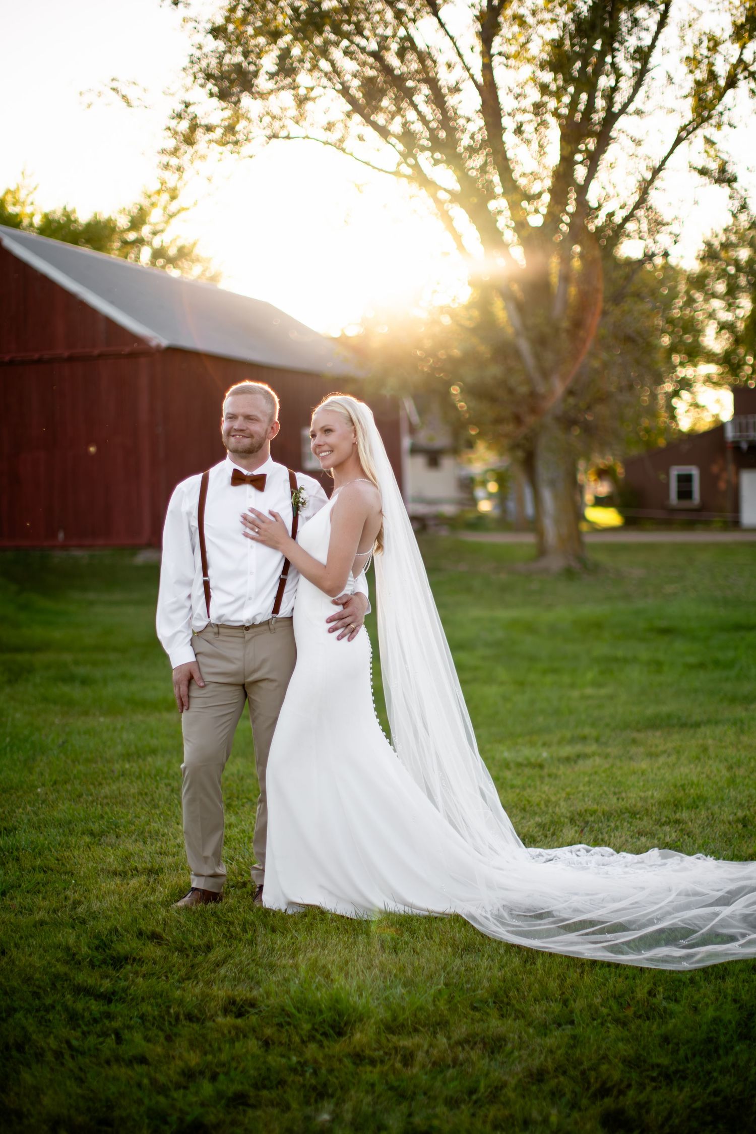 Wedding couple poses by red barn at sunset in rustic countryside setting.