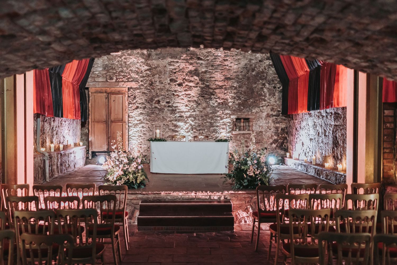 Rustic stone wedding venue with dramatic red lighting and rows of wooden chairs.