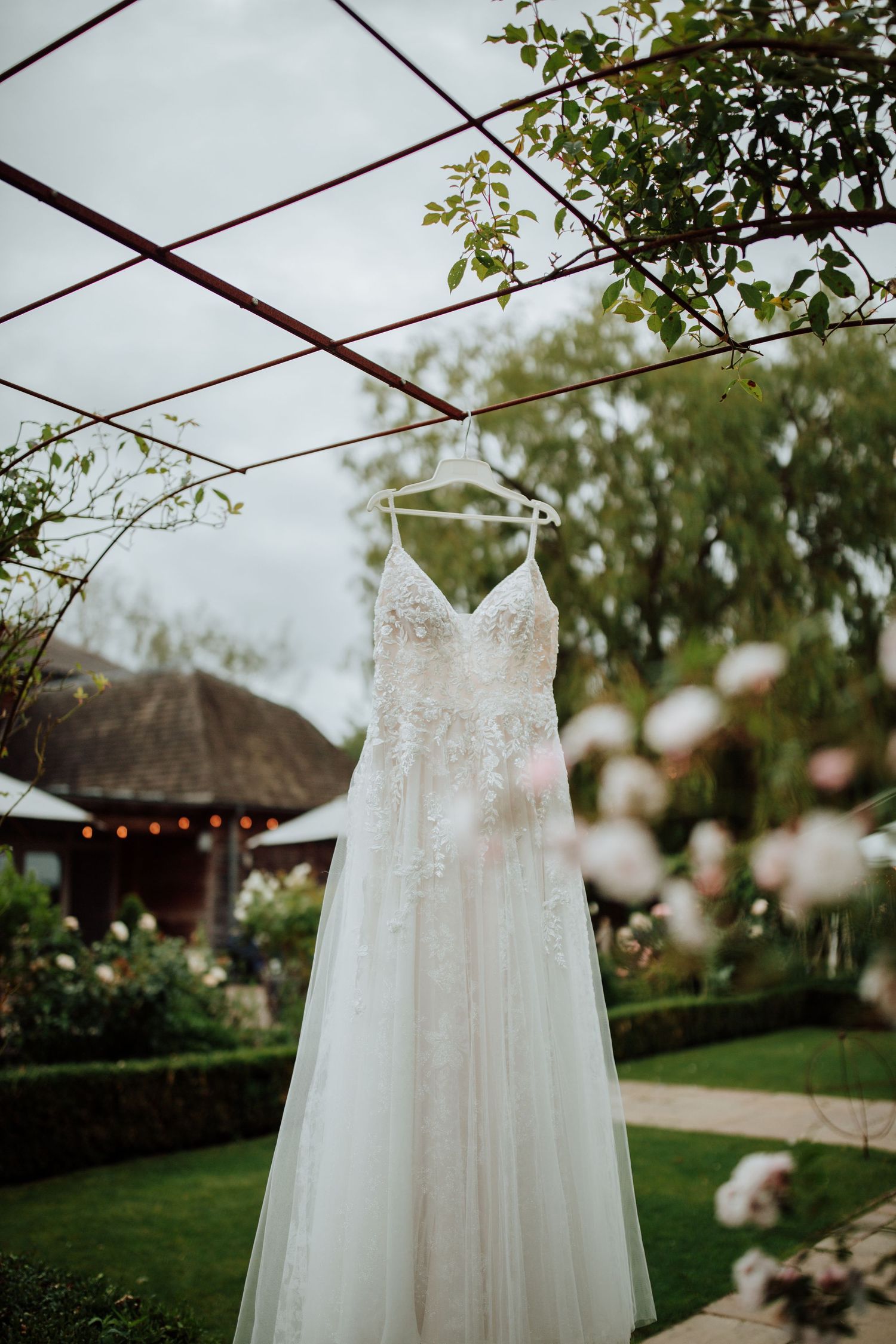 White wedding dress with lace detail hanging in a garden setting with string lights.
