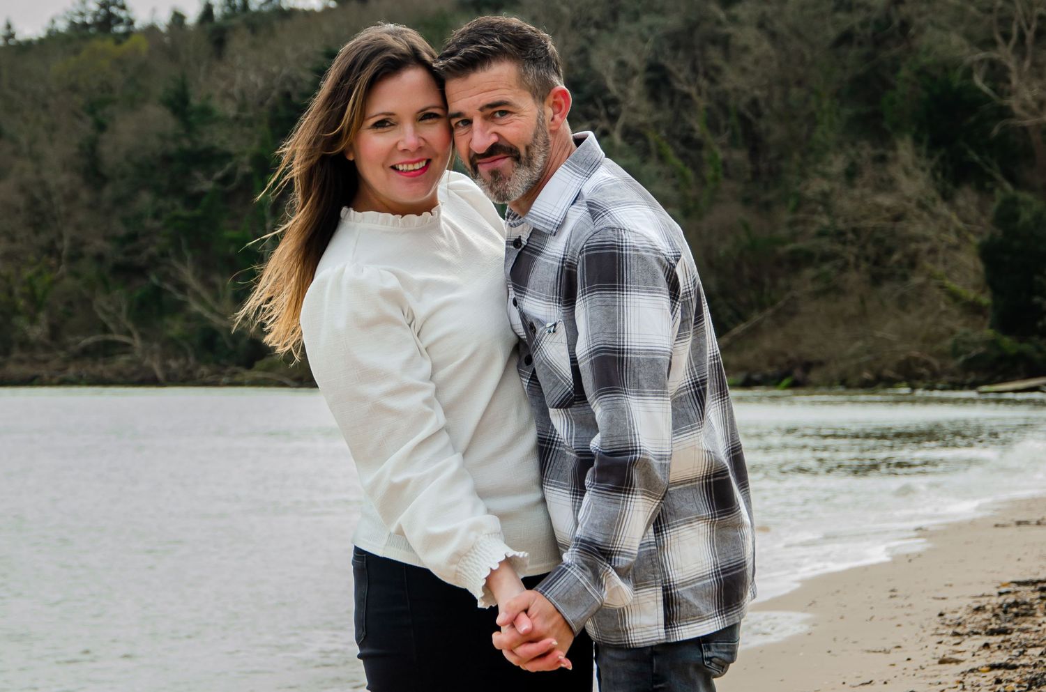 Couple embracing and smiling together on beach with forest background.