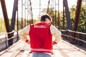 Back view of varsity jacket with text against wooden bridge background.