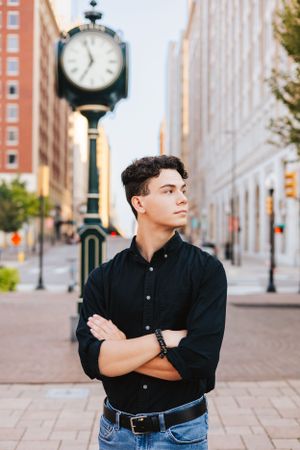 Figure in black button-up shirt poses near vintage street clock in urban setting.