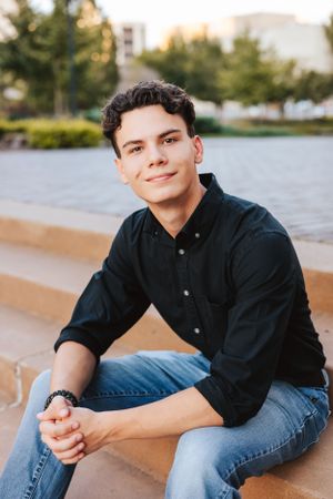 Person in dark shirt and jeans sitting casually by waterfront during sunset.