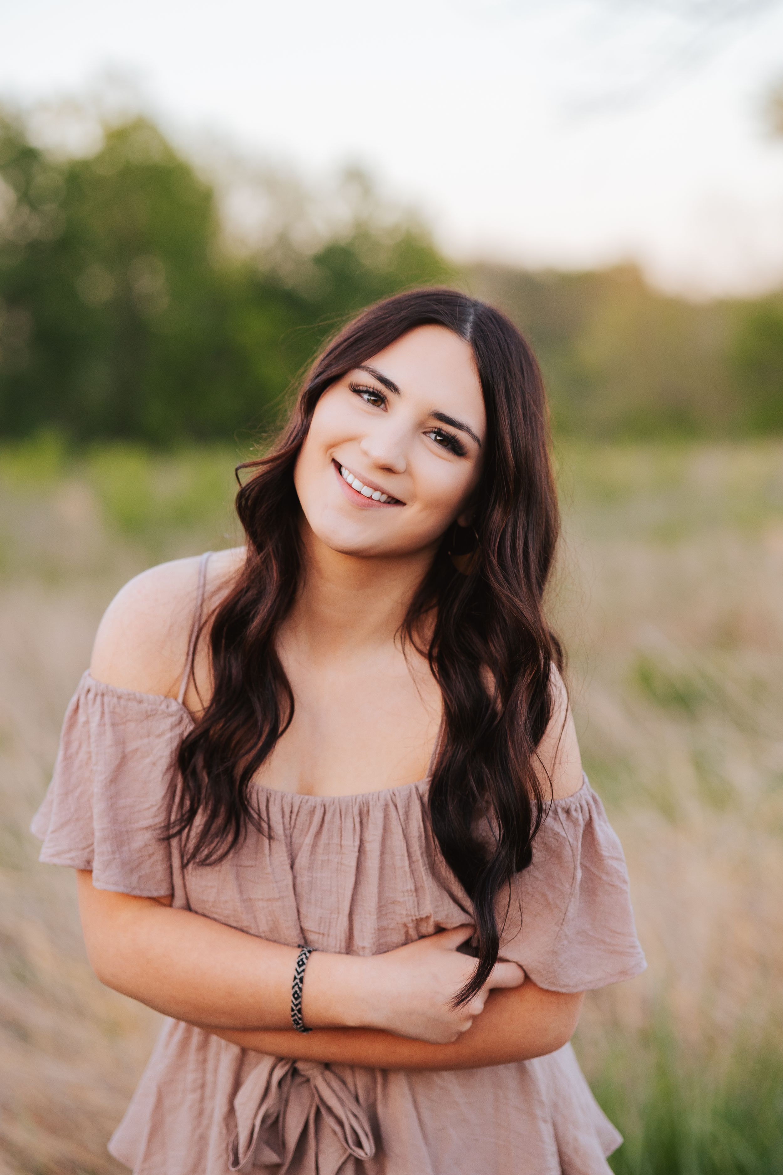Woman in pink off-shoulder dress poses playfully in a grassy field at sunset.
