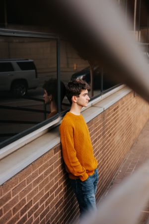 Person in orange sweater and jeans standing against brick wall in artistic portrait series.