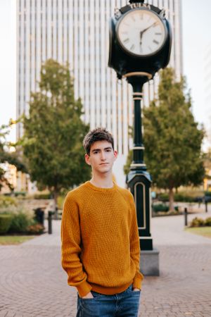 Young man in orange sweater poses near vintage clock in urban setting.