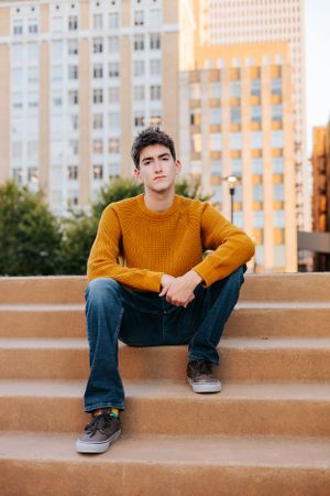 Individual wearing orange sweater sitting on concrete steps with city building in background.