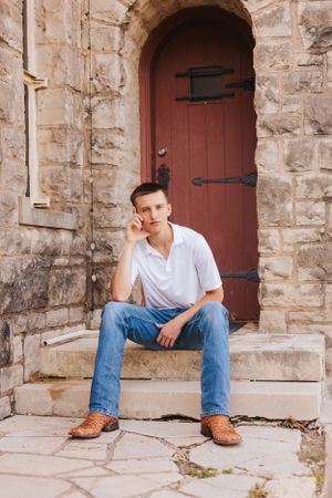 Person in white shirt and jeans sits on stone steps near red door.