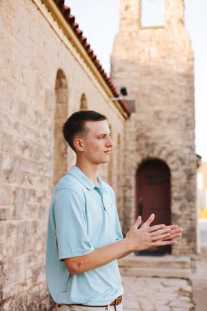 Person in light blue polo gestures while standing by stone building.