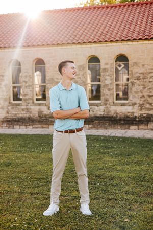 Person in blue polo and khakis stands on grass near stone building.