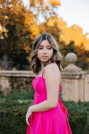 A bright pink formal dress flows elegantly against an autumn backdrop with stone walls and golden foliage.