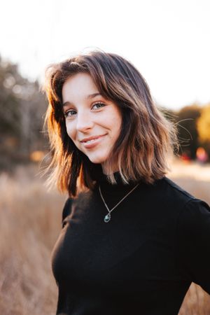 Woman with short brown hair smiles warmly in black turtleneck at sunset.