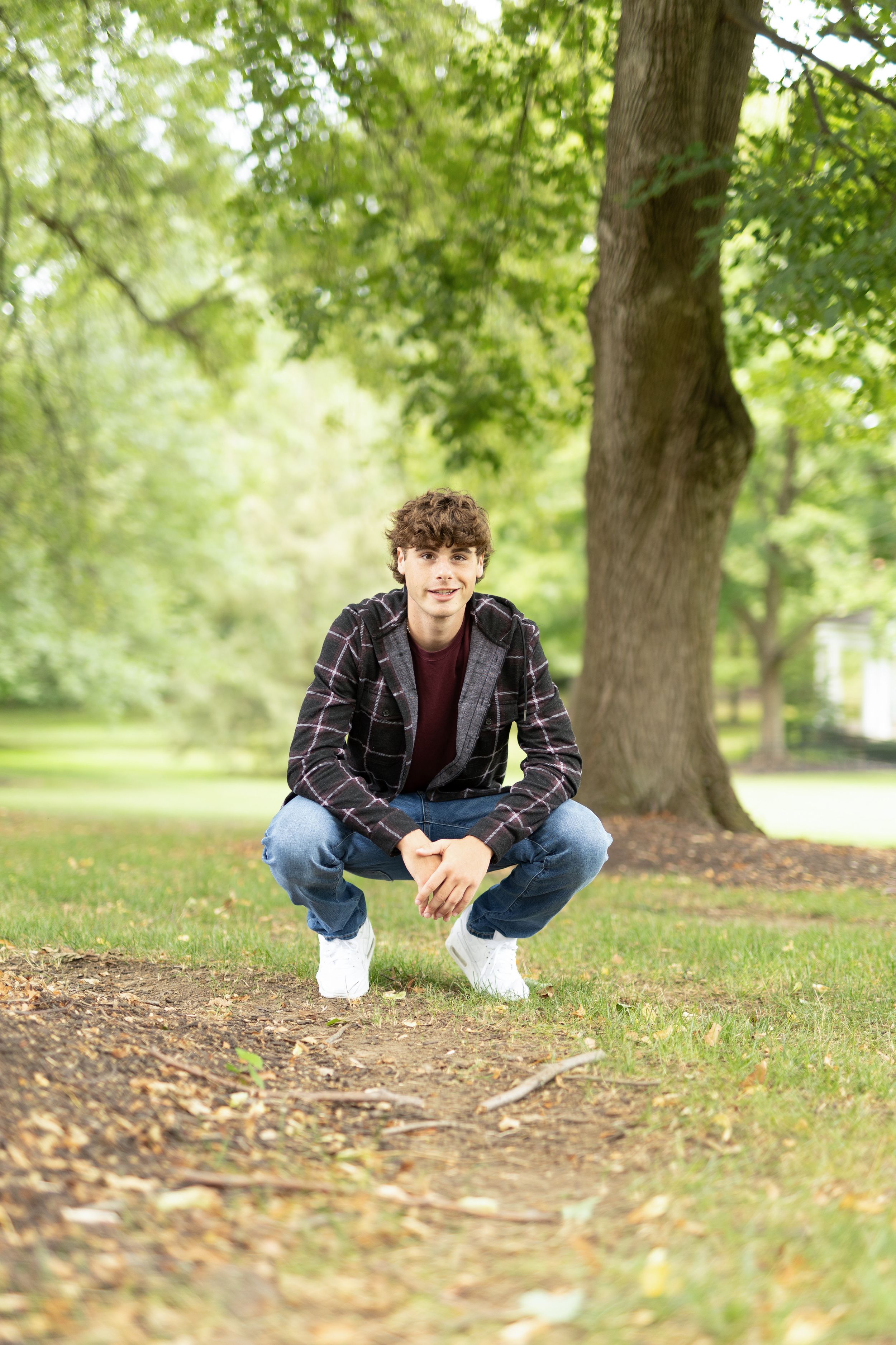A guy crouching in a park setting surrounded by fallen leaves and trees.