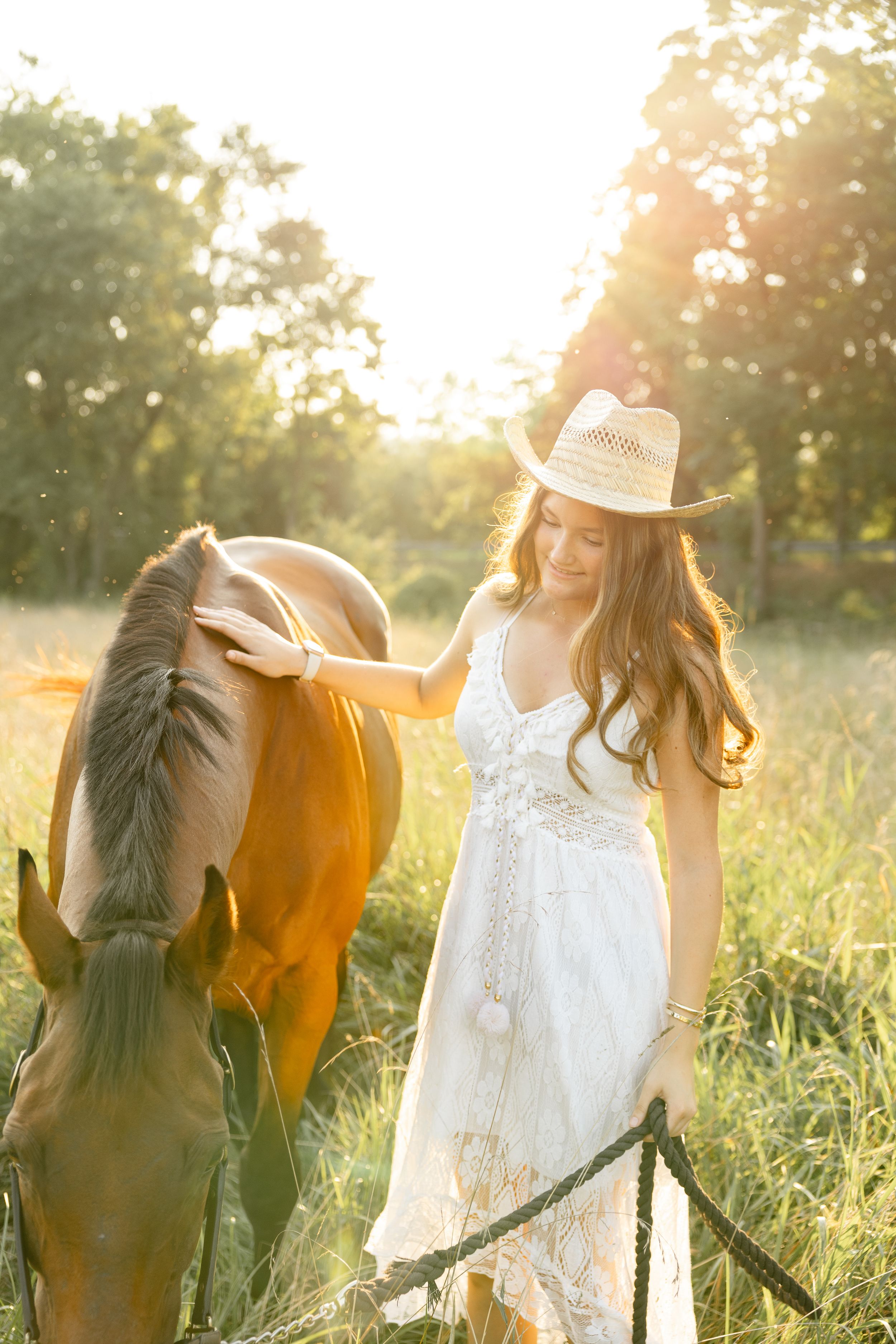 A person in a white dress and straw hat stands with a brown horse in a sunlit meadow during golden hour.