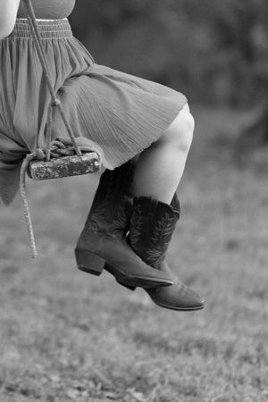 A close-up of cowboy boots and a flowing dress captured in black and white, sitting on a wooden swing.