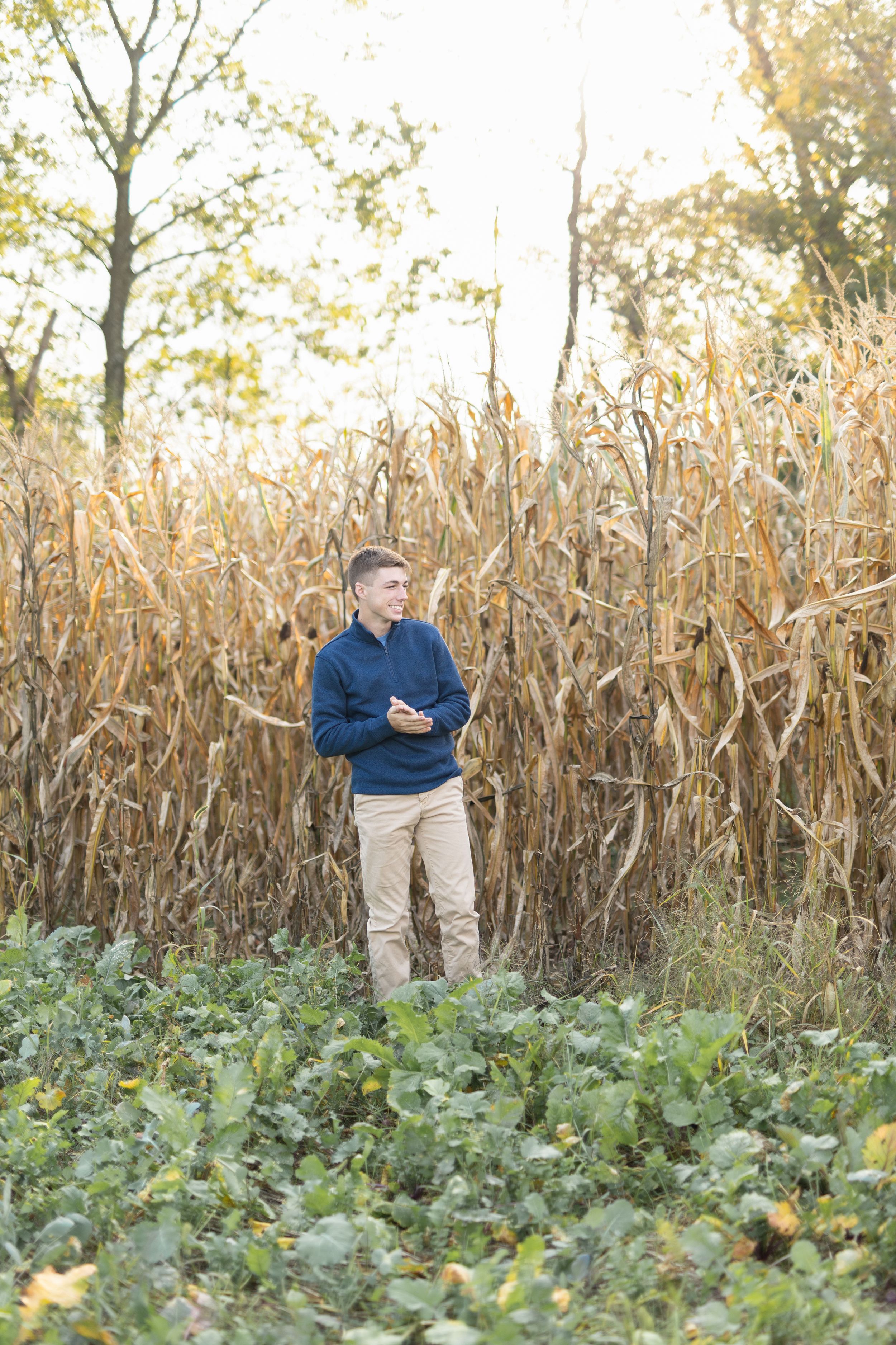 A guy in a navy sweater stands among tall corn stalks in an autumn field.