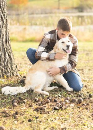 A fluffy dog its owner posing in autumn leaves while sitting at the base of a tree.