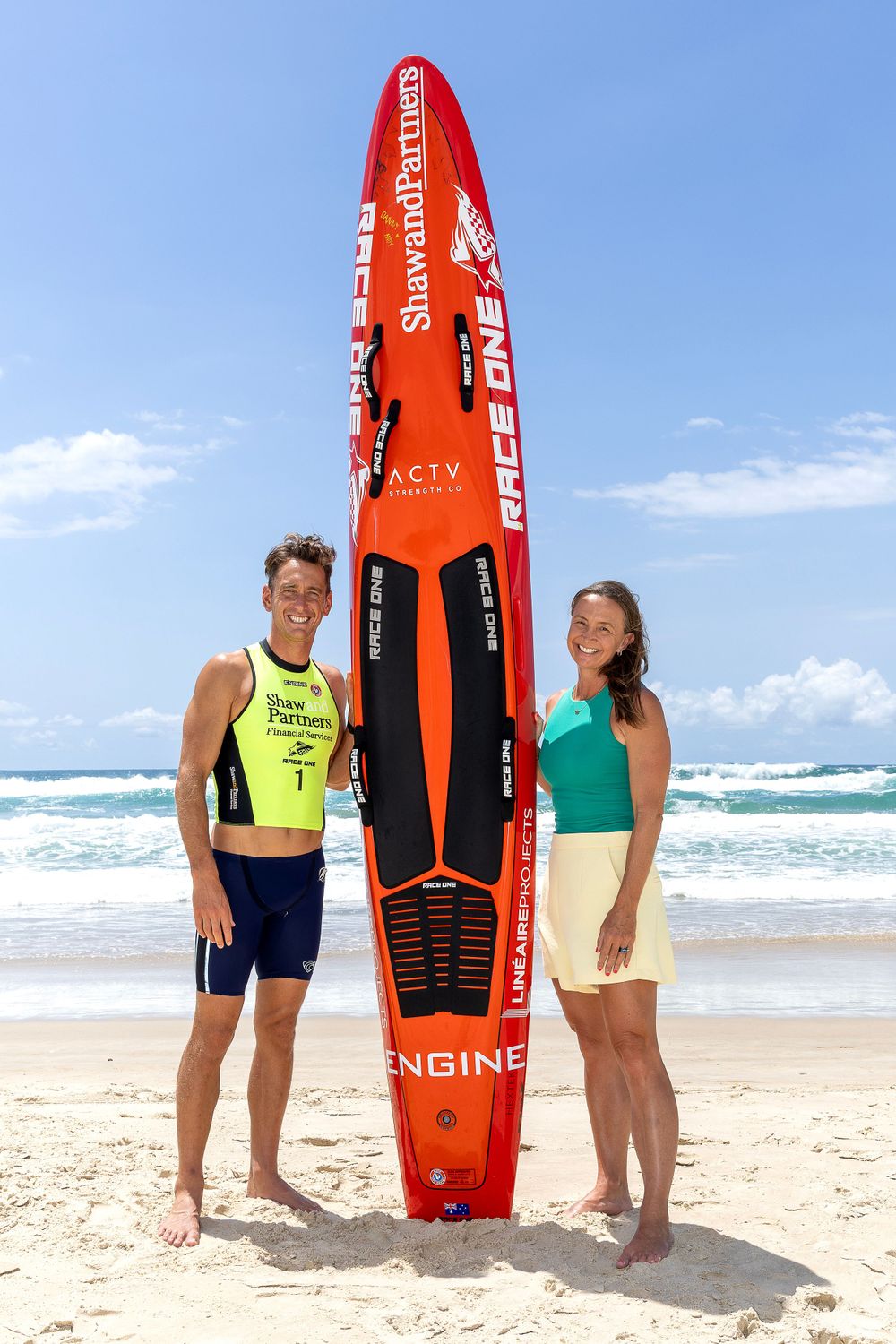 Athletes standing with orange racing surfboard on sandy beach with waves in background.