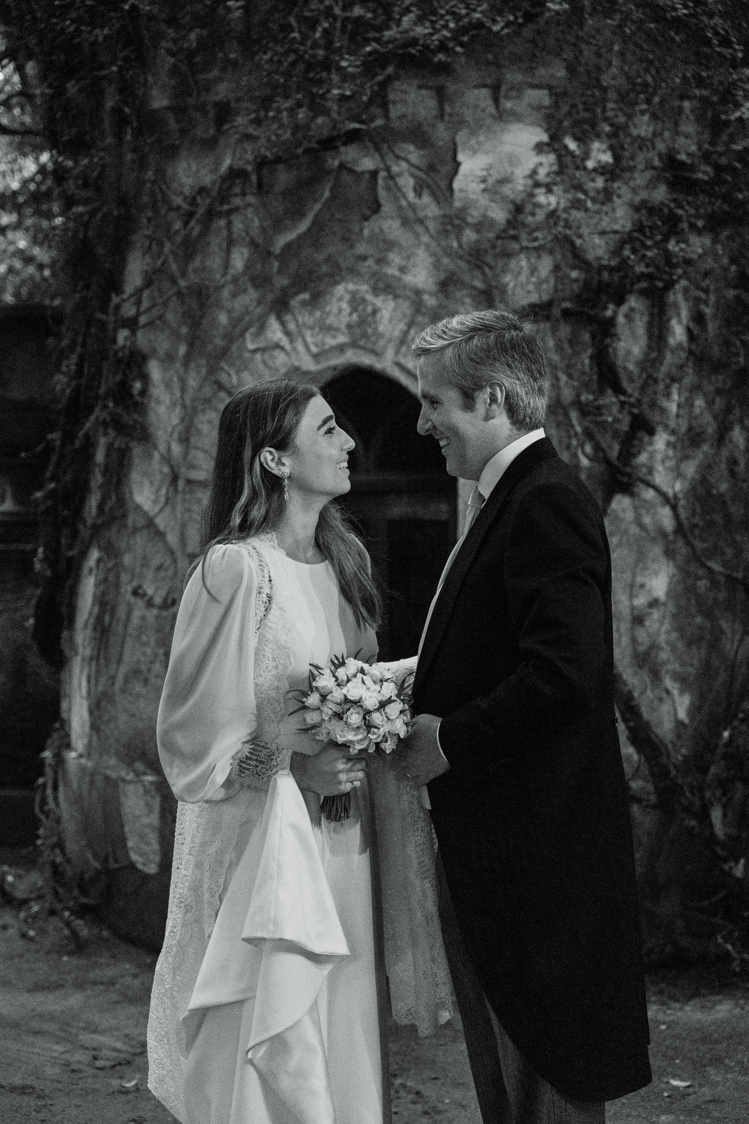 Fotografia de casamento mostrando um casal de noivos a olhar-se com amor diante de uma antiga estrutura de pedra coberta de hera.