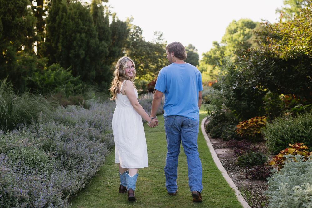 A person in a white dress and another in jeans walk along a garden path surrounded by blooming flowers.