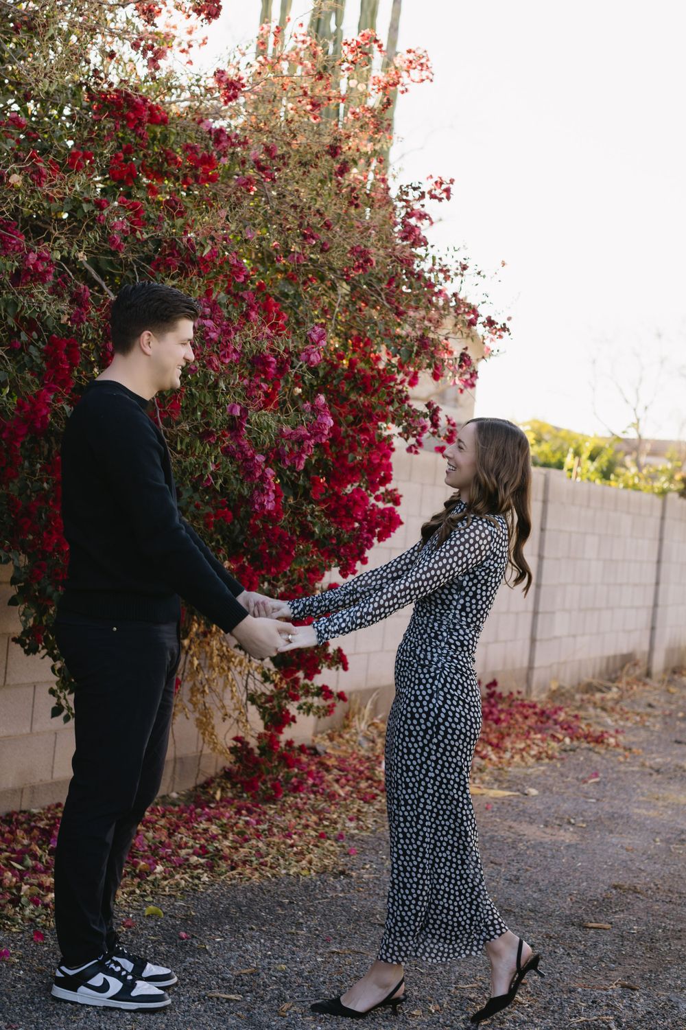 Two people hold hands while standing in front of vibrant red bougainvillea flowers against a block wall.