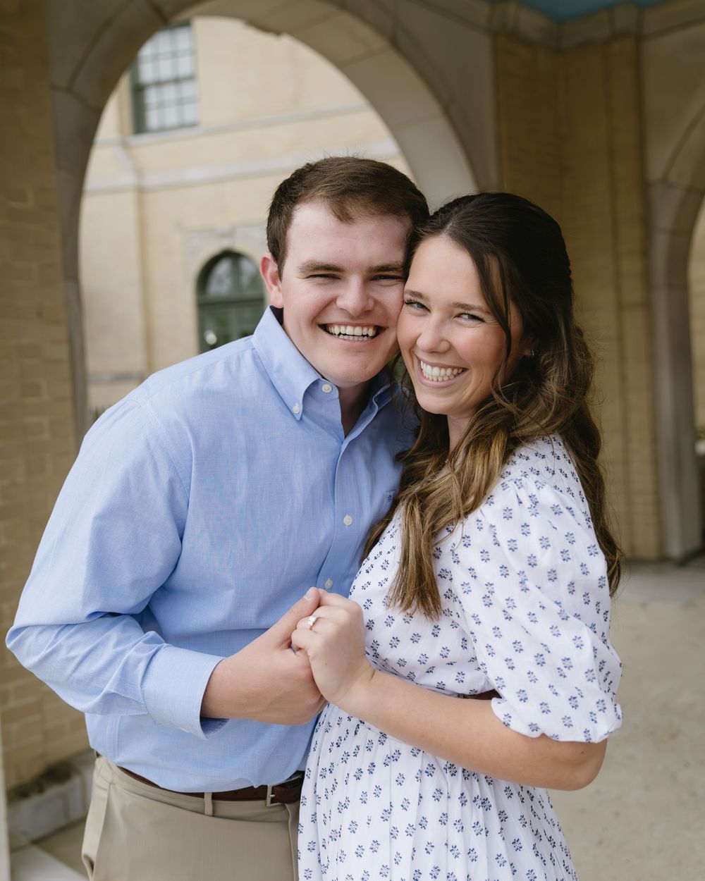 A couple poses together and smiles beneath an arched walkway, wearing a blue button-down shirt and floral dress.