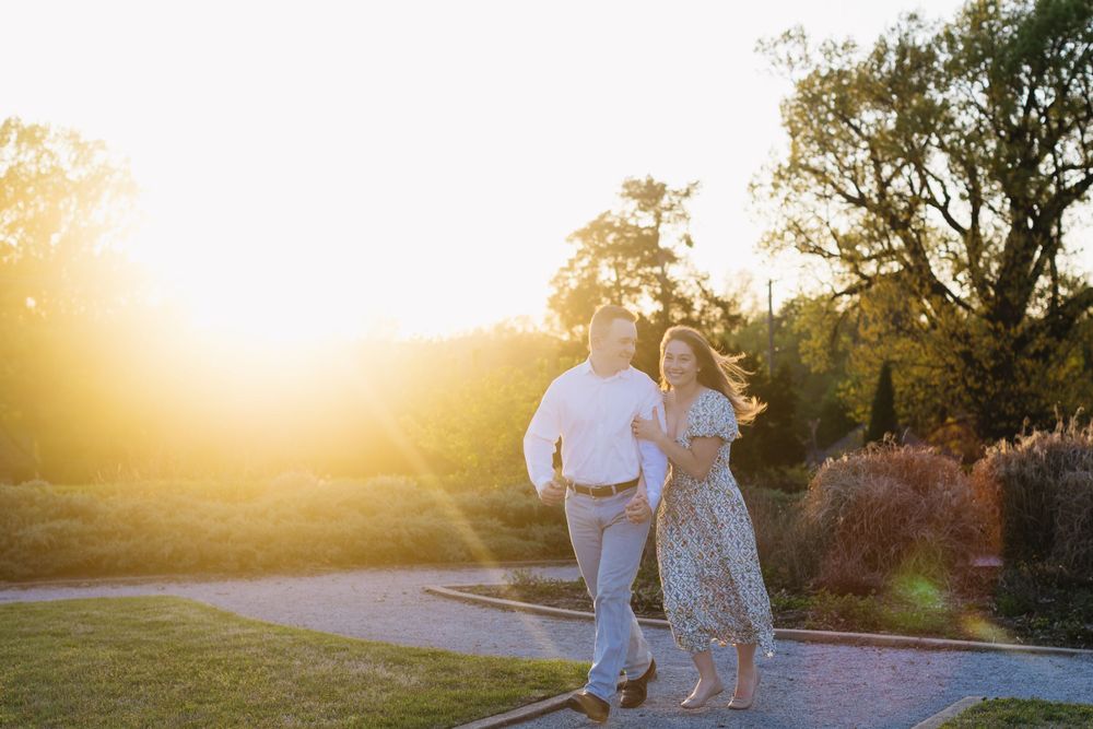 Two people walk along a path during golden hour sunset with sunlight streaming through trees in a park setting.