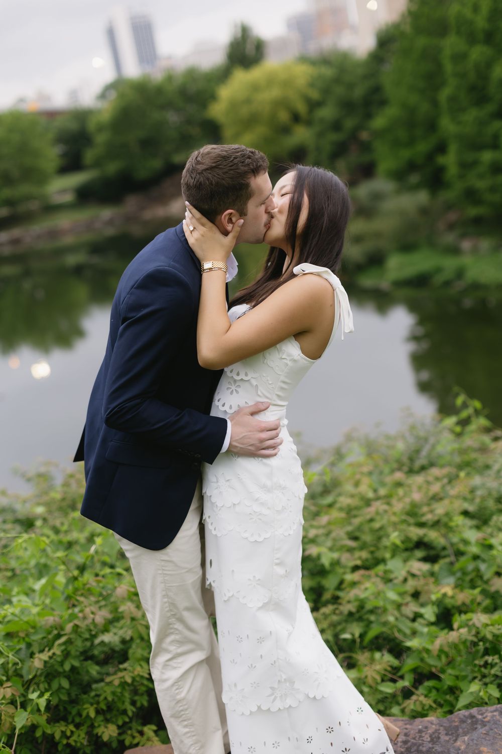 A couple shares a romantic kiss in an outdoor garden setting with city skyline visible in background.