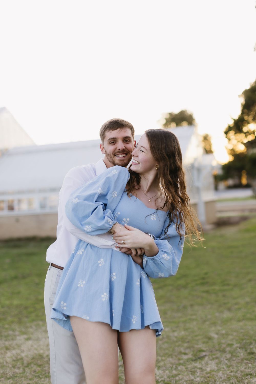 A romantic couple embraces outdoors in matching blue outfits during golden hour with a white barn in the background.