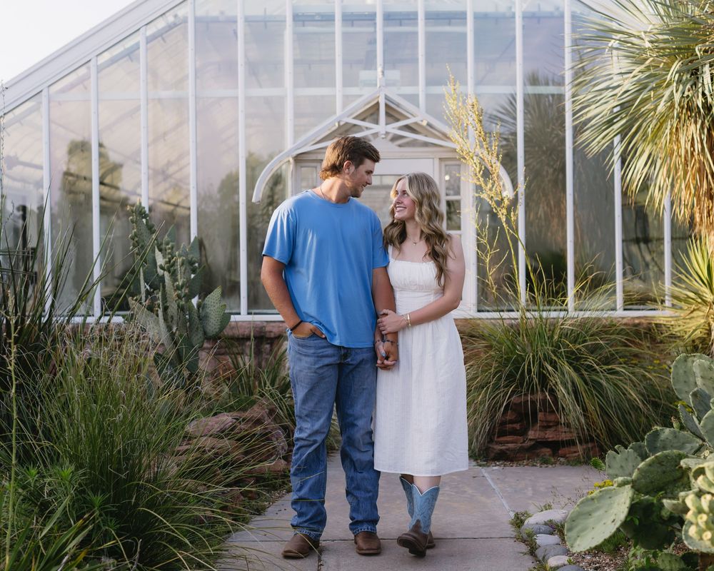 A couple in casual clothing shares intimate moments together in a dreamy greenhouse setting with palm trees.