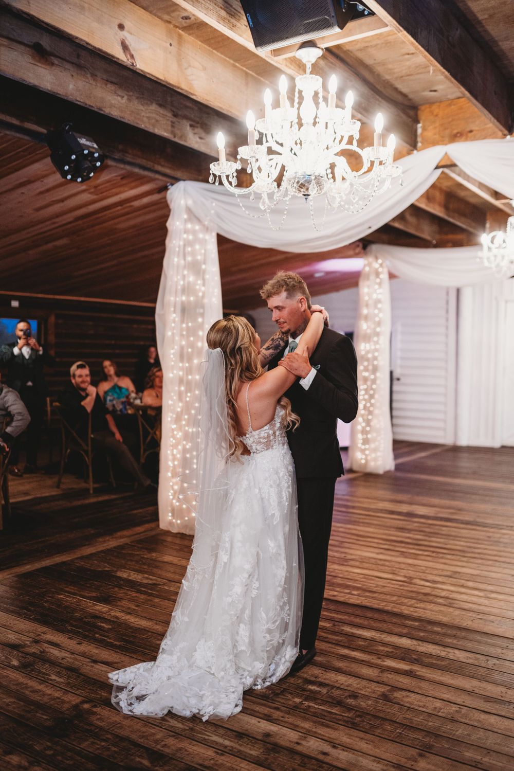 First dance under crystal chandelier in rustic wooden venue with white draped decor.