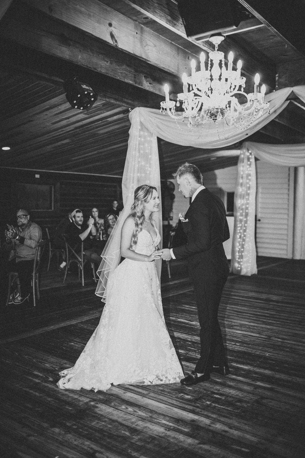A couple shares their first dance under an elegant crystal chandelier at their rustic wedding reception.