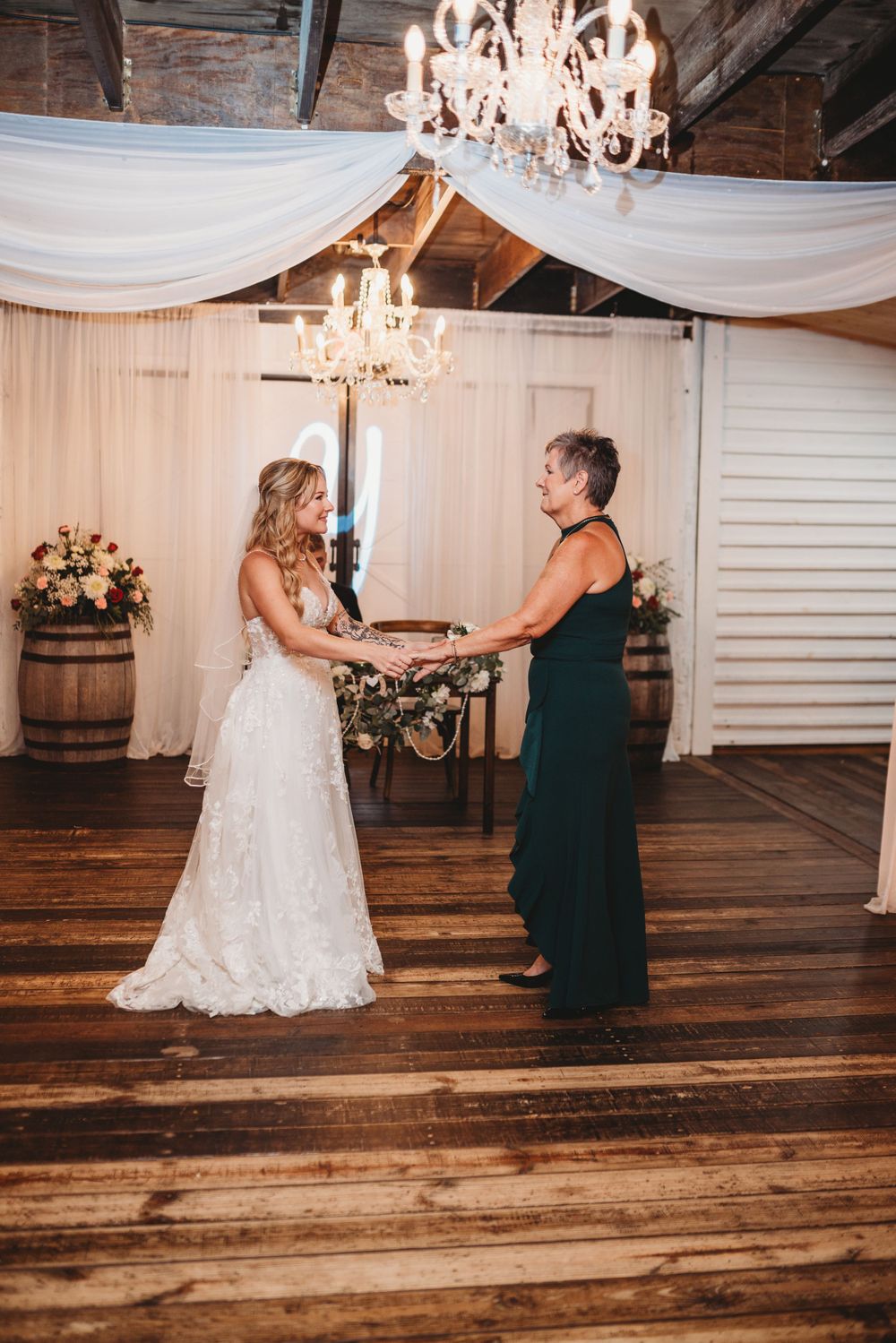 A bride in a white lace gown shares a tender moment with a guest under draped fabric and chandeliers.